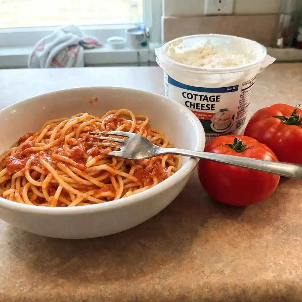 A bowl of high protein red sauce pasta beside cottage cheese and tomatoes showing its nutrition