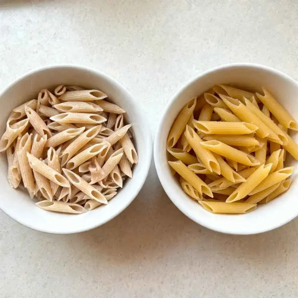 Protein penne pasta next to regular penne in two bowls, comparing color and texture